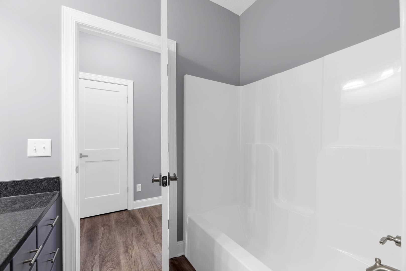 White freestanding bathtub beside wood plank flooring, white door with silver handle, dual light switch on plaster wall, countertop visible in foreground