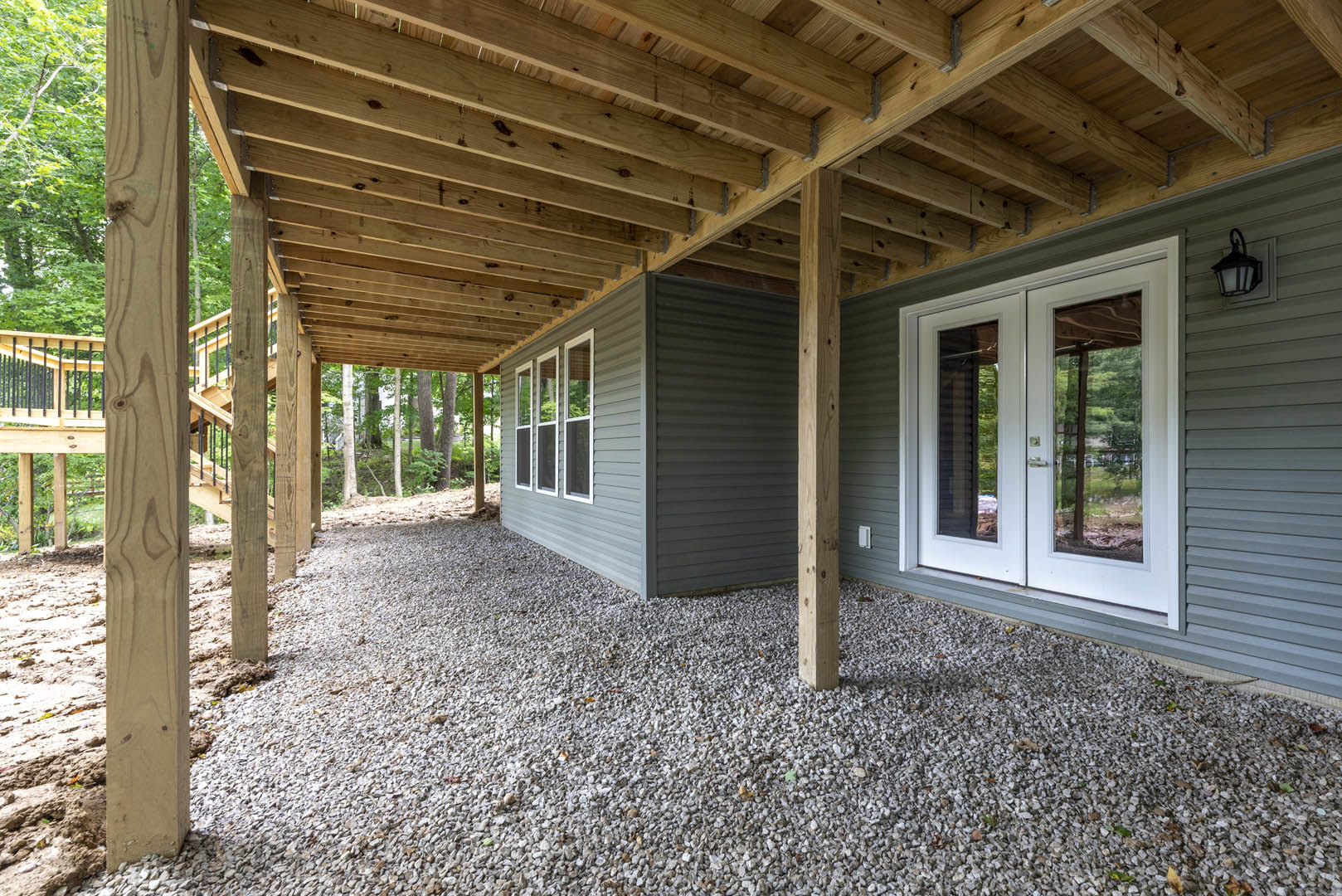 Covered patio with wooden beams and posts, double glass doors, gravel ground, wall-mounted light fixture, window overlooking trees.