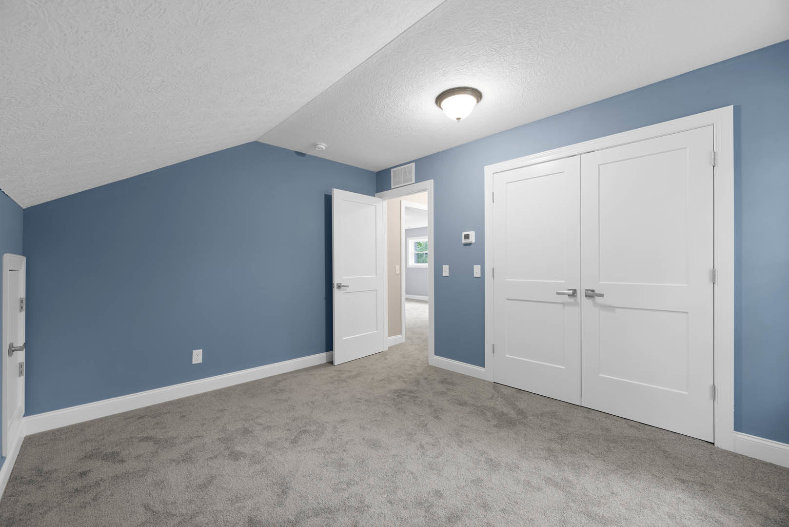 Carpeted room with blue accent wall, two white doors featuring silver handles, ceiling light fixture, and wall vent
