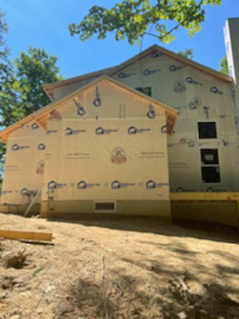 Partially built house with white weatherproof covering, exposed window opening, dirt mound in foreground, leafy tree branches and wooded area in background