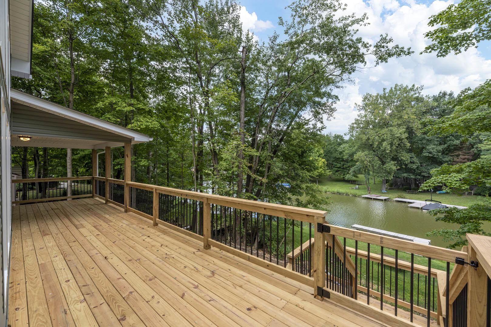 Wooden deck with black metal railing, surrounded by green trees, overlooking a calm lake under a partly cloudy sky