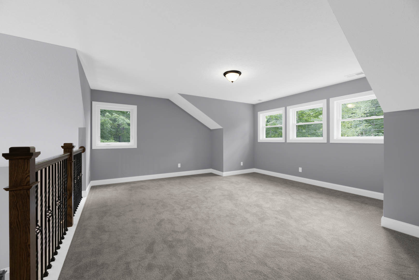 Grey carpeted room with white walls, multiple windows showing trees outside, ceiling light fixture, close-up of stair railing, white trim and molding