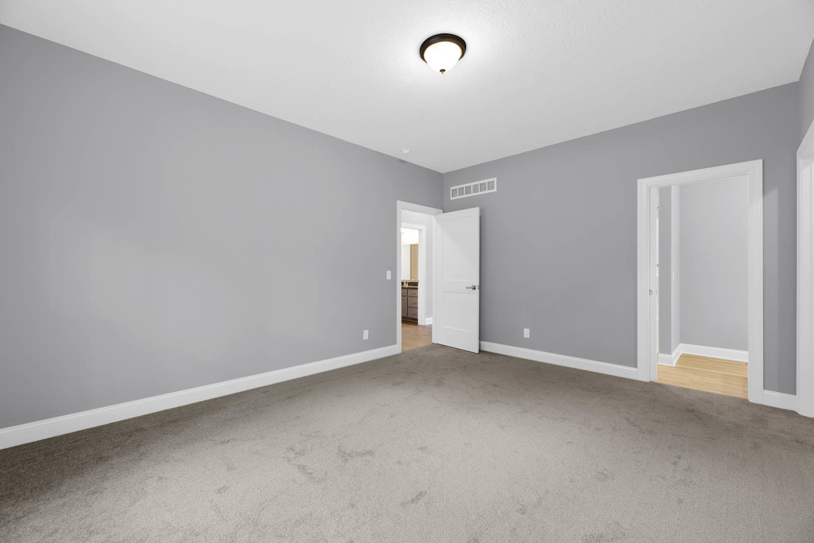 Carpeted room with white walls, white door with silver handle and white frame, ceiling light fixture, white wall vent, plaster molding along ceiling.