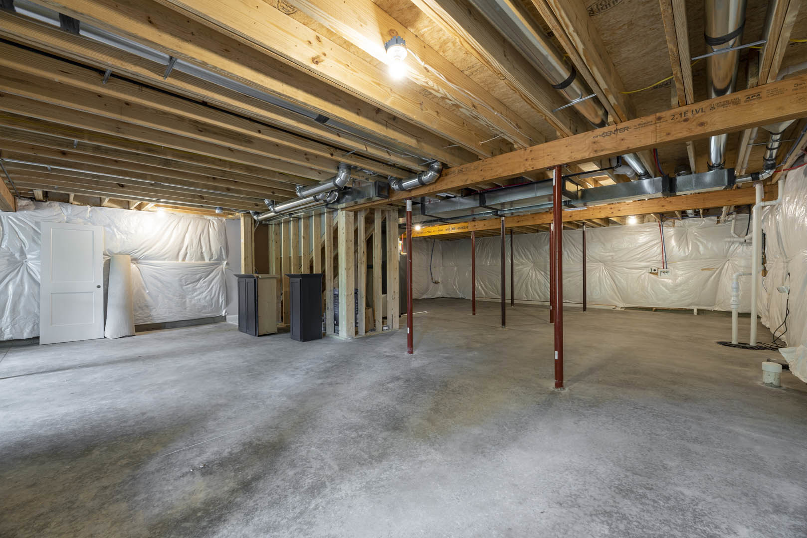 Exposed wood ceiling beams and metal pipes above unfinished room, white door with doorknob, white sheet draped along wall, light fixture illuminated, concrete floor