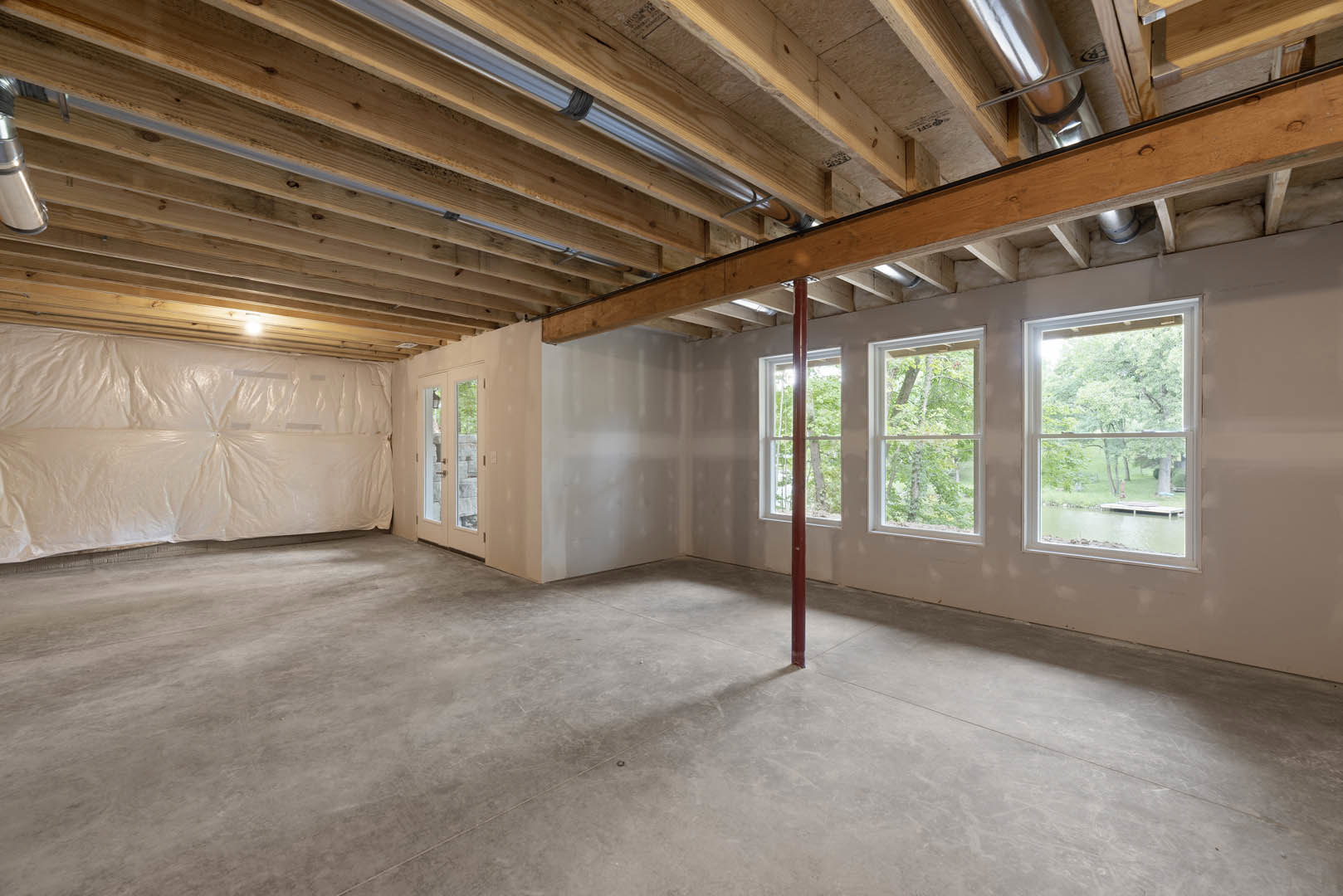 Room with exposed wooden ceiling, large windows overlooking trees and lake, concrete floor with red support pole, white plastic covering part of wall.