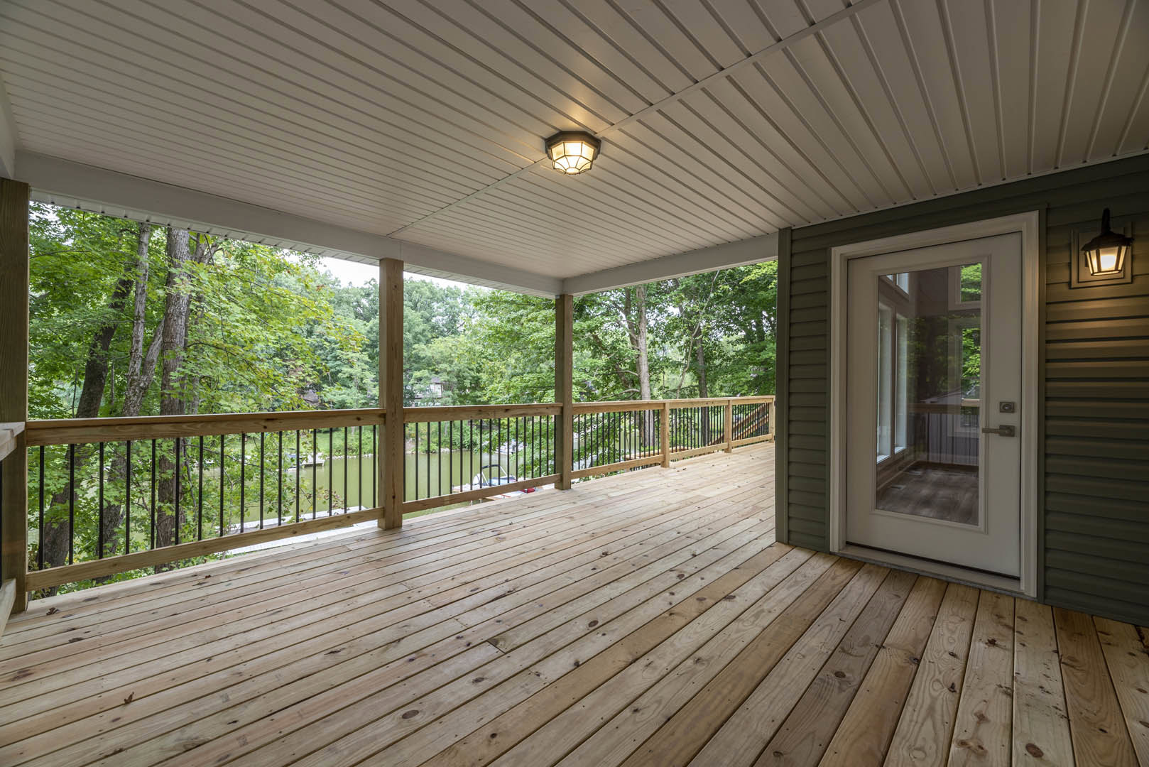 Wooden deck with metal railing, glass door leading inside, ceiling light fixture, lake and trees visible in background
