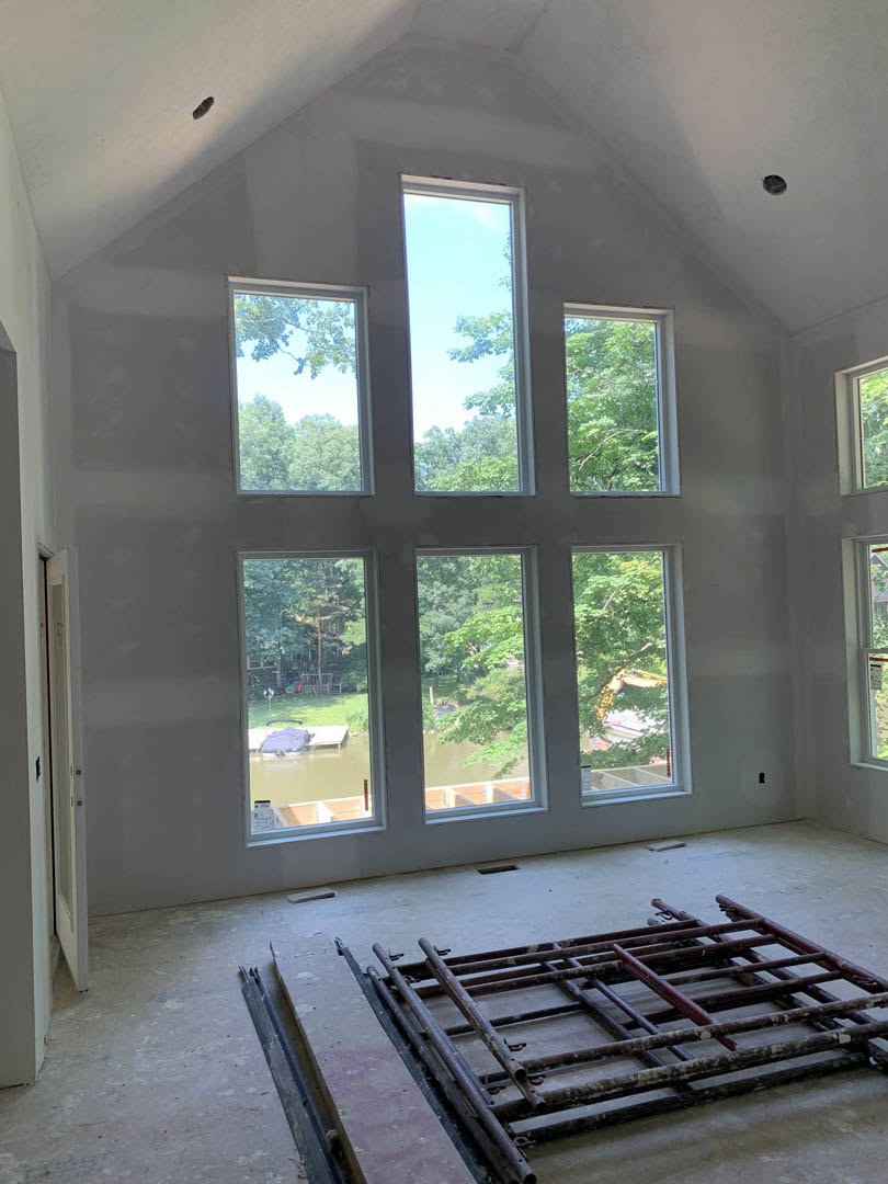 Spacious room featuring multiple large windows framed in wood, light streaming in, metal frame resting on hardwood floor, trees visible outside through glass panes, white walls and