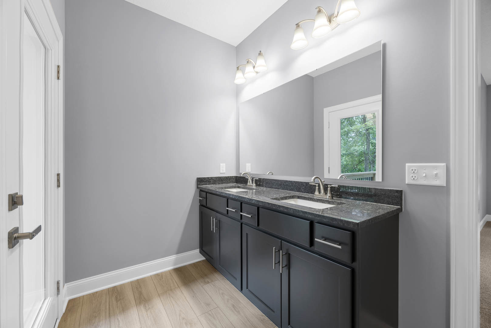 Bathroom with black cabinets, large mirror above white countertop, modern light fixture, wooden floor with white baseboard, window showing trees outside, close-up of door and wall
