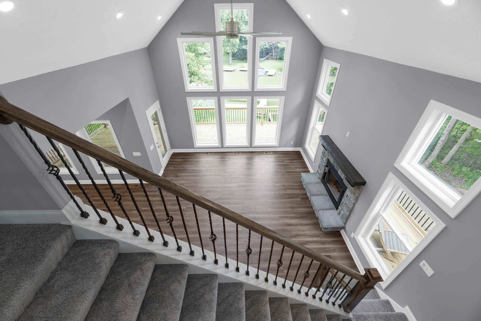 Staircase with grey carpet and wooden handrail descending to wooden floor, black-framed fireplace set in white wall, large window overlooking backyard with trees, ceiling fan above