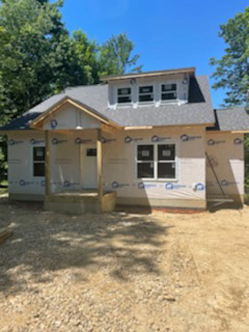 Framed house under construction with exposed wooden beams, partially installed roof, surrounded by mature trees and blue sky