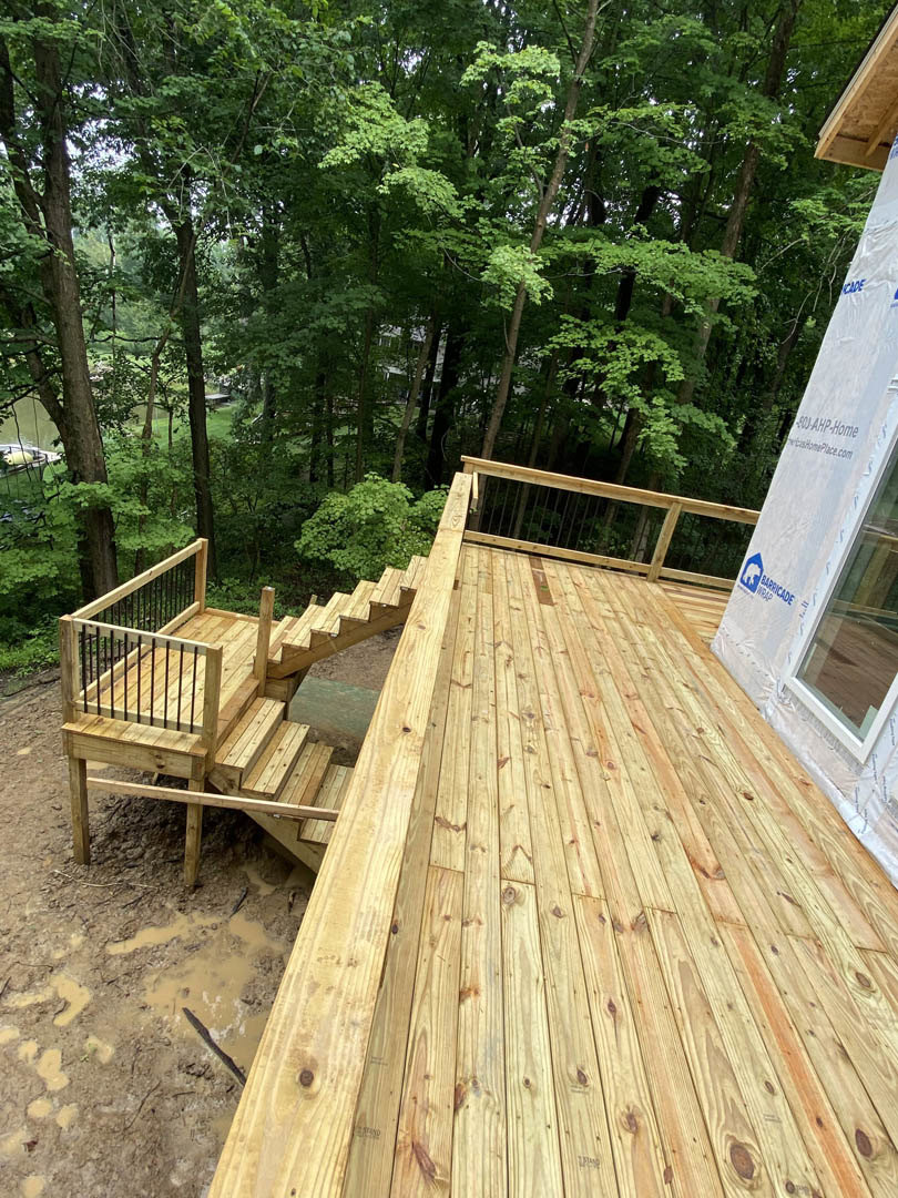Wooden deck with black metal railings and stairs surrounded by dense trees, muddy ground beneath, white sign with black text attached to railing