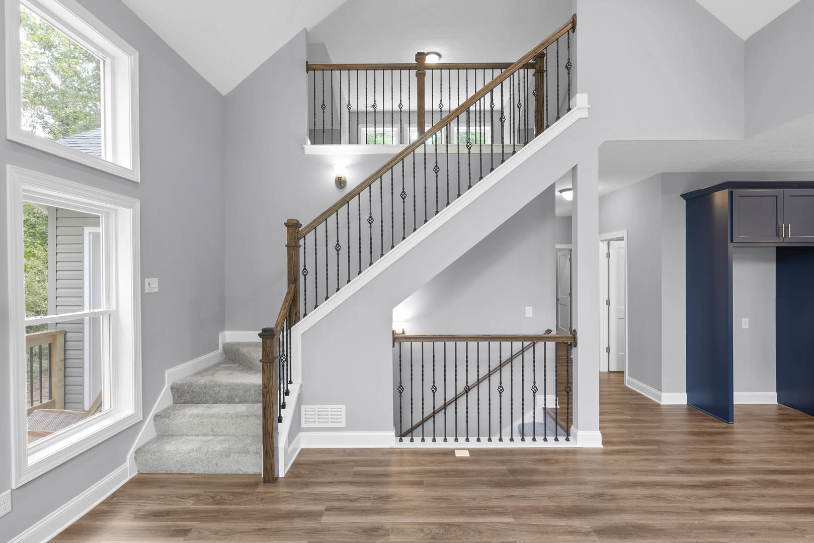 Wood staircase with black metal railings, white vent on wall, wide plank wood flooring, large window revealing leafy tree outside
