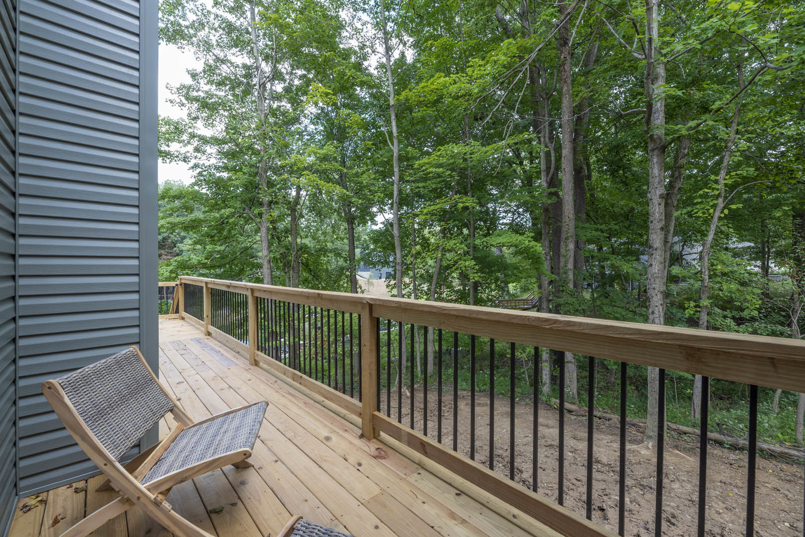 Wooden deck with black railings, outdoor chairs, and mature trees in the background; close-up of grey exterior wall visible.