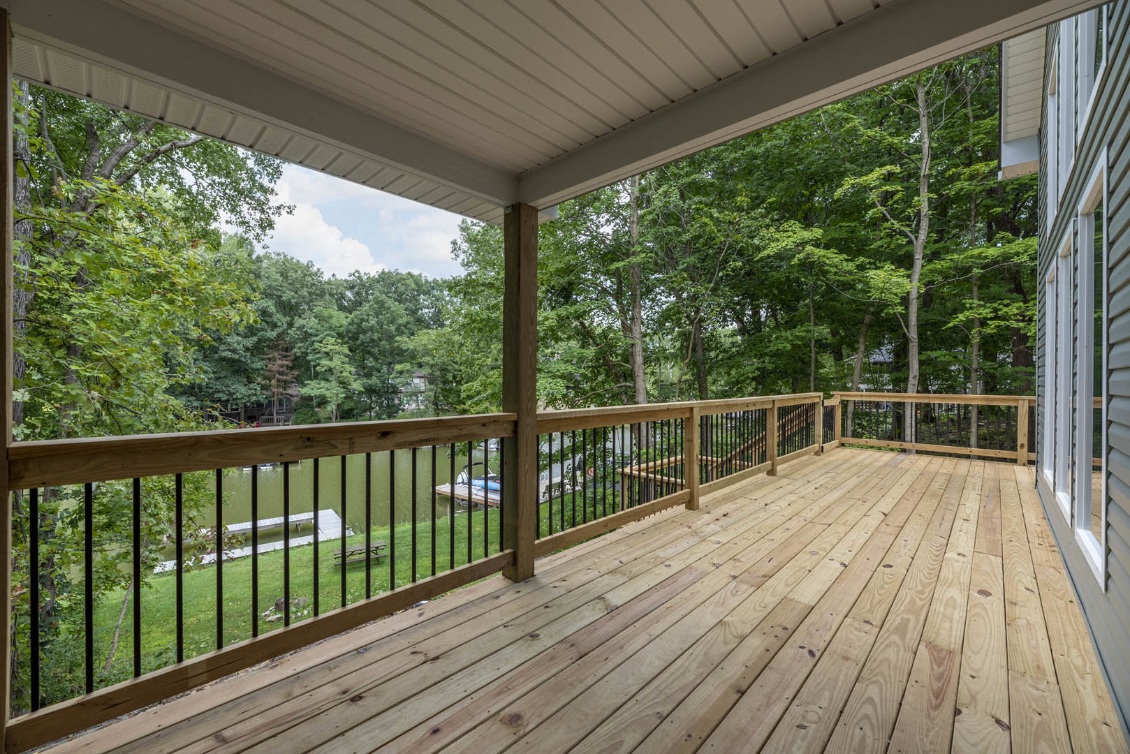 Wooden deck with railing overlooking grassy yard and mature trees, lake visible in background, bench near pond, white ceiling and roof above.