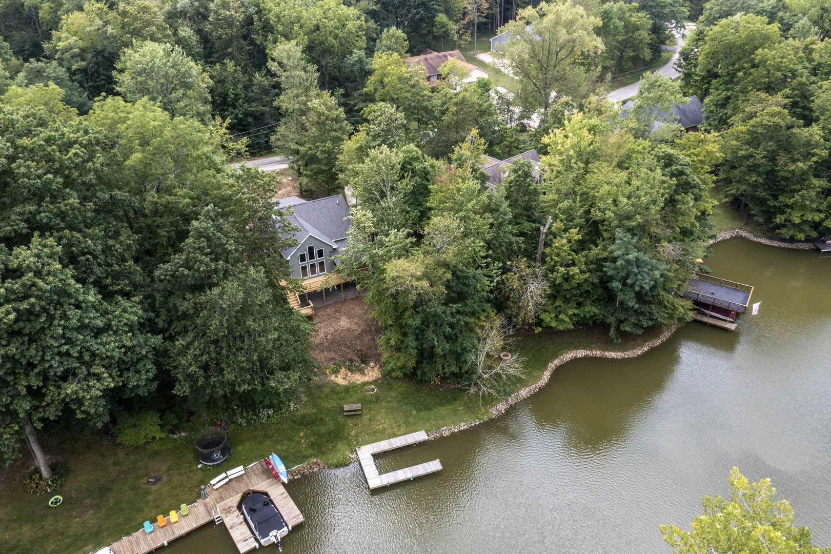 Two-story house with light siding and large windows, surrounded by mature trees, situated beside a calm lake with a wooden dock extending into the water; small boat moored at the