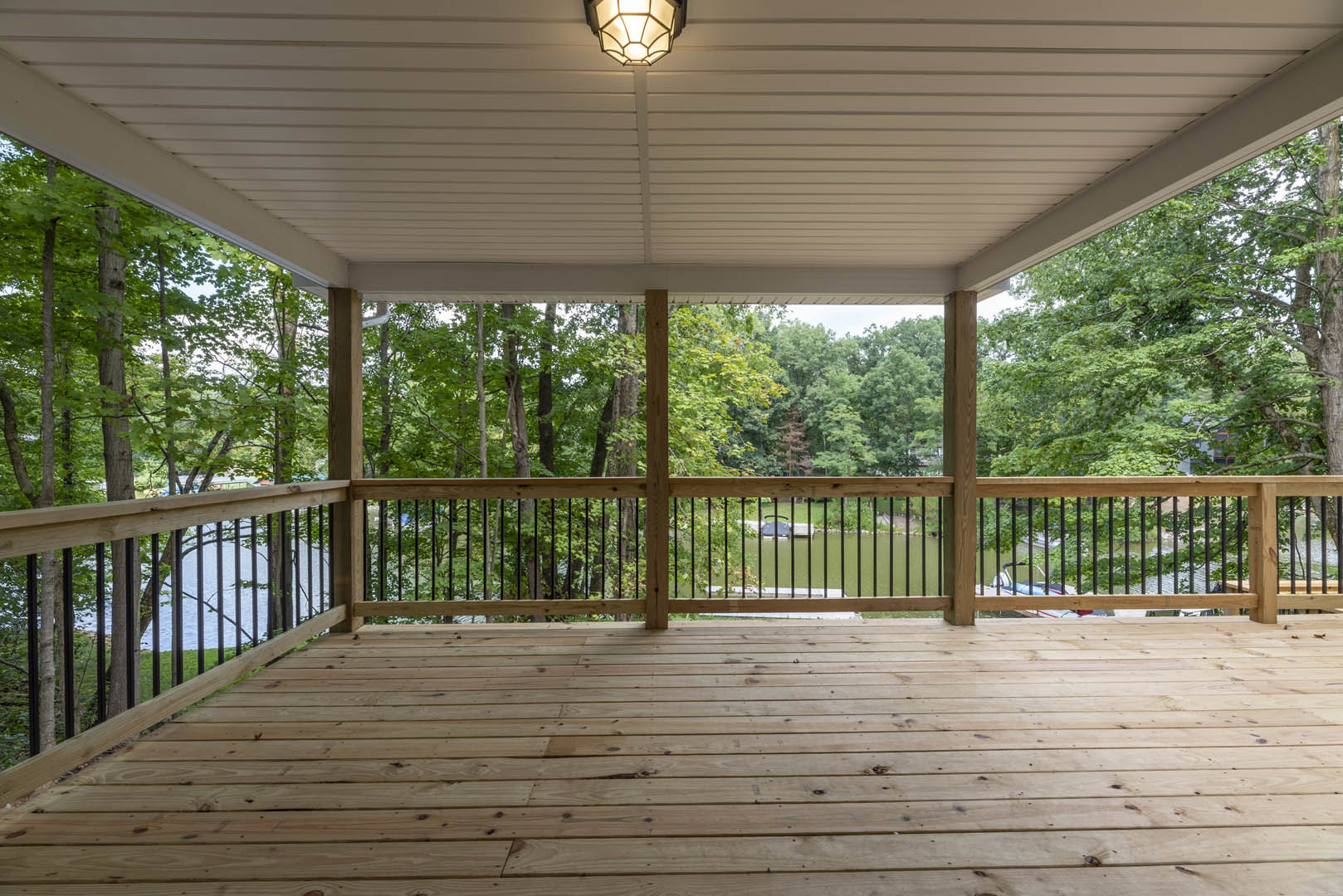 Wooden deck with black metal railing, overhead light fixture, surrounded by leafy trees and partial water view