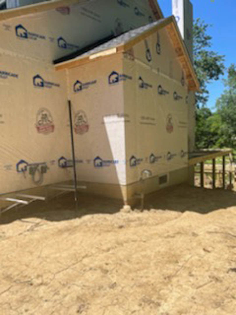 Partially built home with completed roof, exposed framing, and visible insulation, surrounded by dirt lot and construction materials.