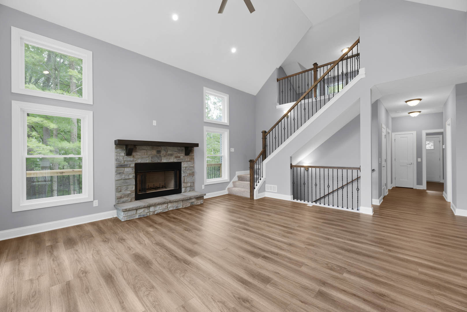 Living room with hardwood floors, modern fireplace featuring a glass door and wood mantel, large window overlooking trees, staircase with metal railings.