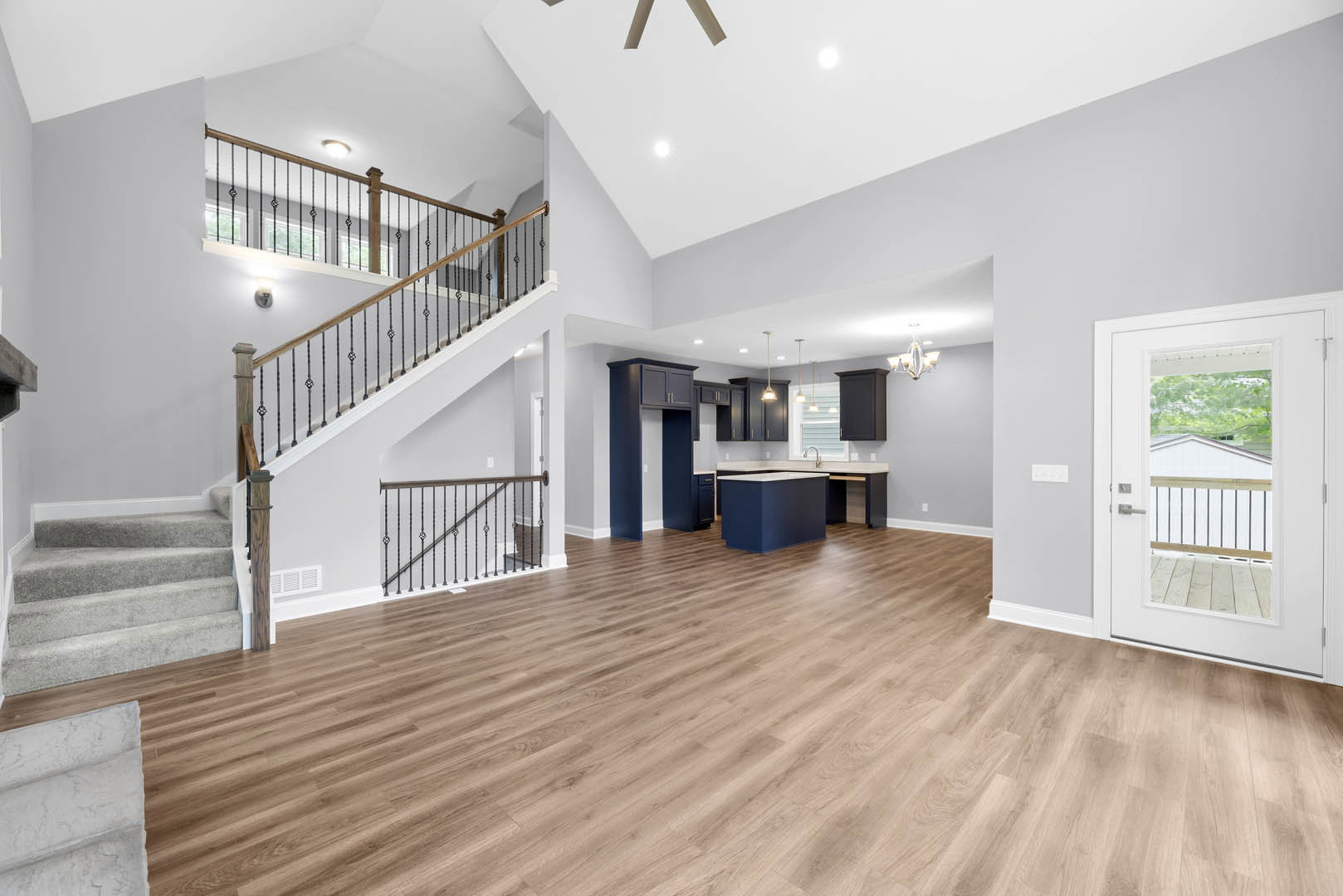 Open-concept room featuring hardwood floors, a modern metal-railed staircase, white walls, a kitchen area with a blue island and white countertop, and a glass-paneled white door.