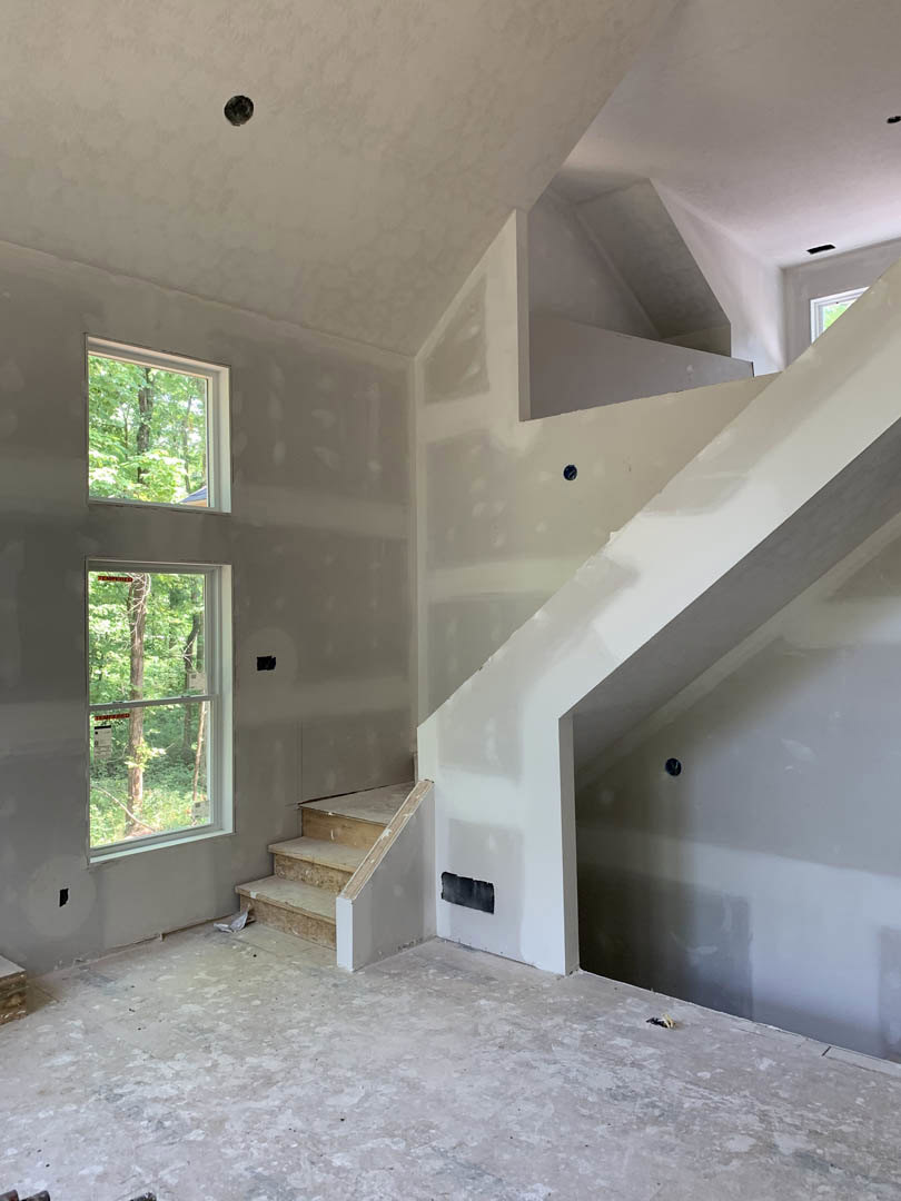 White-walled room featuring a modern white staircase, large windows with white frames, and light wood flooring; trees visible outside through the windows.