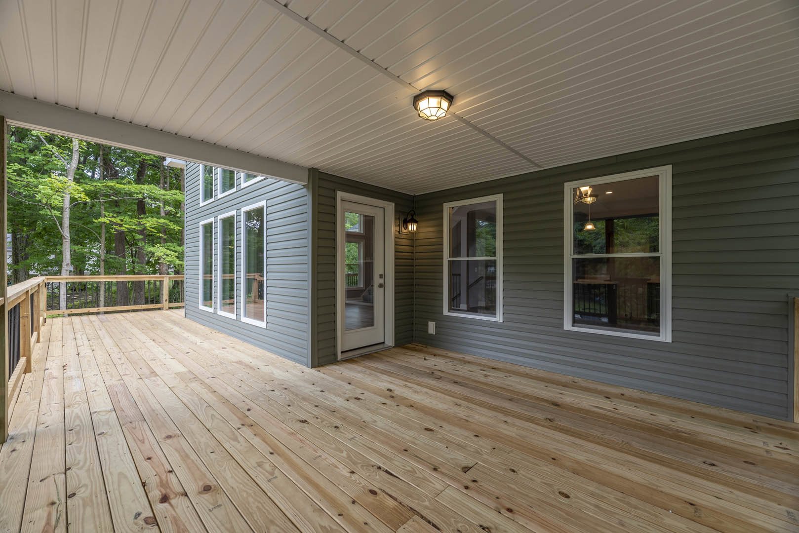 Wood deck with natural plank flooring, covered porch area, white framed windows, glass door, ceiling fan, and overhead light fixture; leafy trees visible through windows.