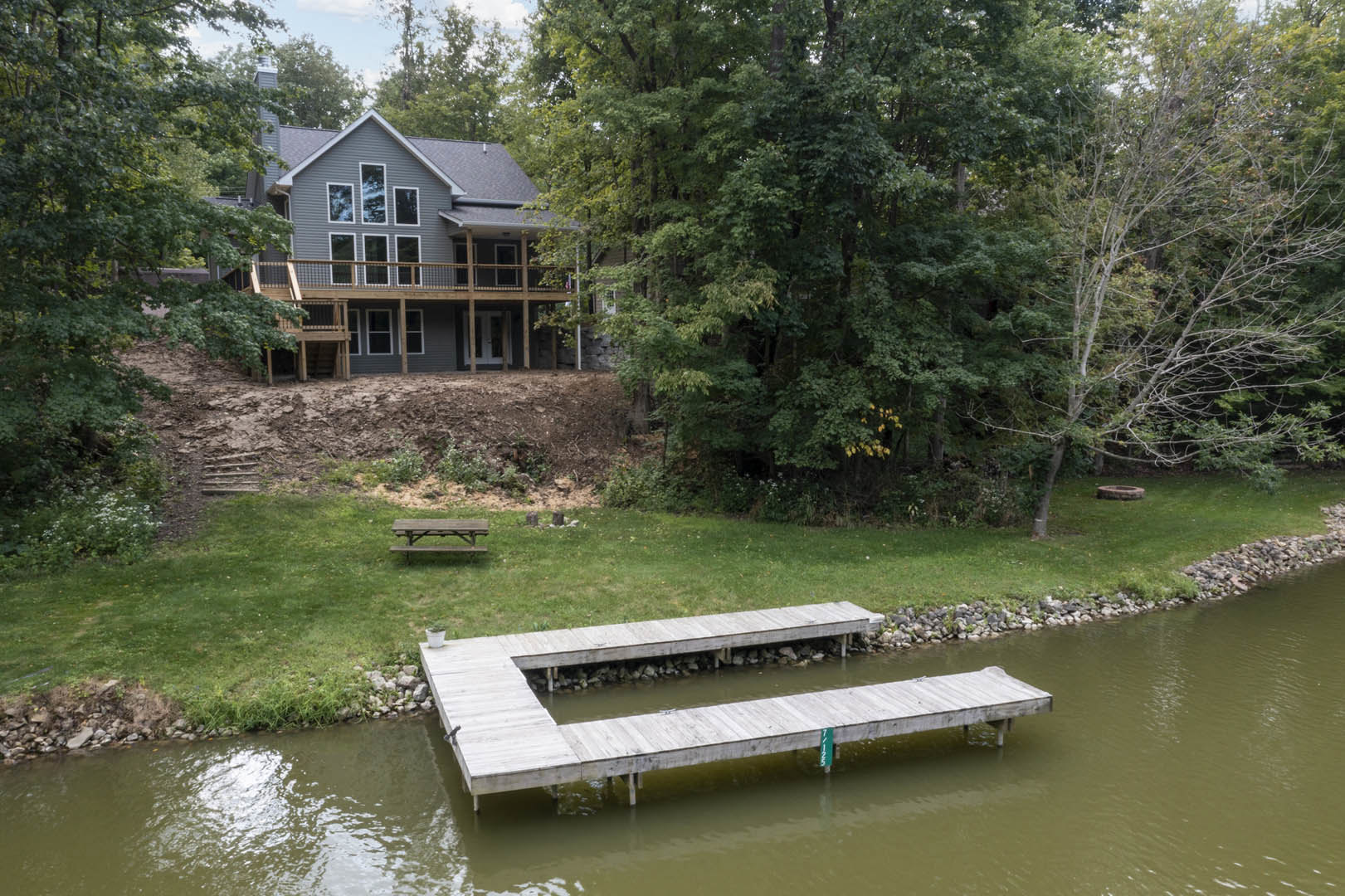 Two-story house with white siding, upper balcony, and ground-level deck overlooking a wooden dock extending over calm water, surrounded by green grass, mature trees, and blue sky.