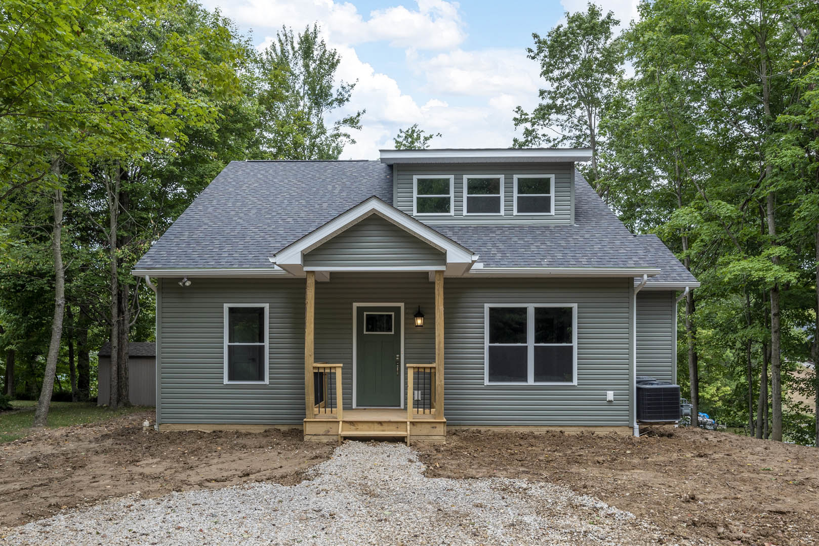 Front porch with wooden steps, green door with window, white-framed windows, driveway bordered by trees, light-colored siding