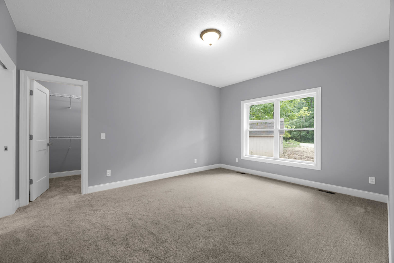 Carpeted room with a large window overlooking trees, white door with silver handle, ceiling light fixture, and wall vent
