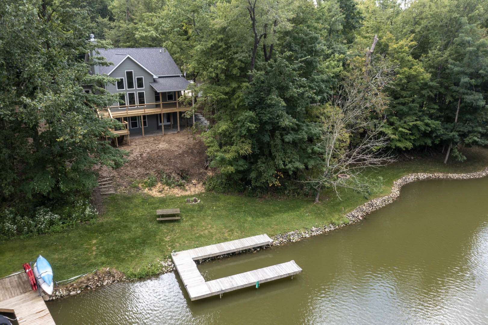 Two-story house with gray siding and white trim, elevated deck overlooking a grassy yard, wooden dock extending into calm lake, blue and white kayak beside dock, mature trees