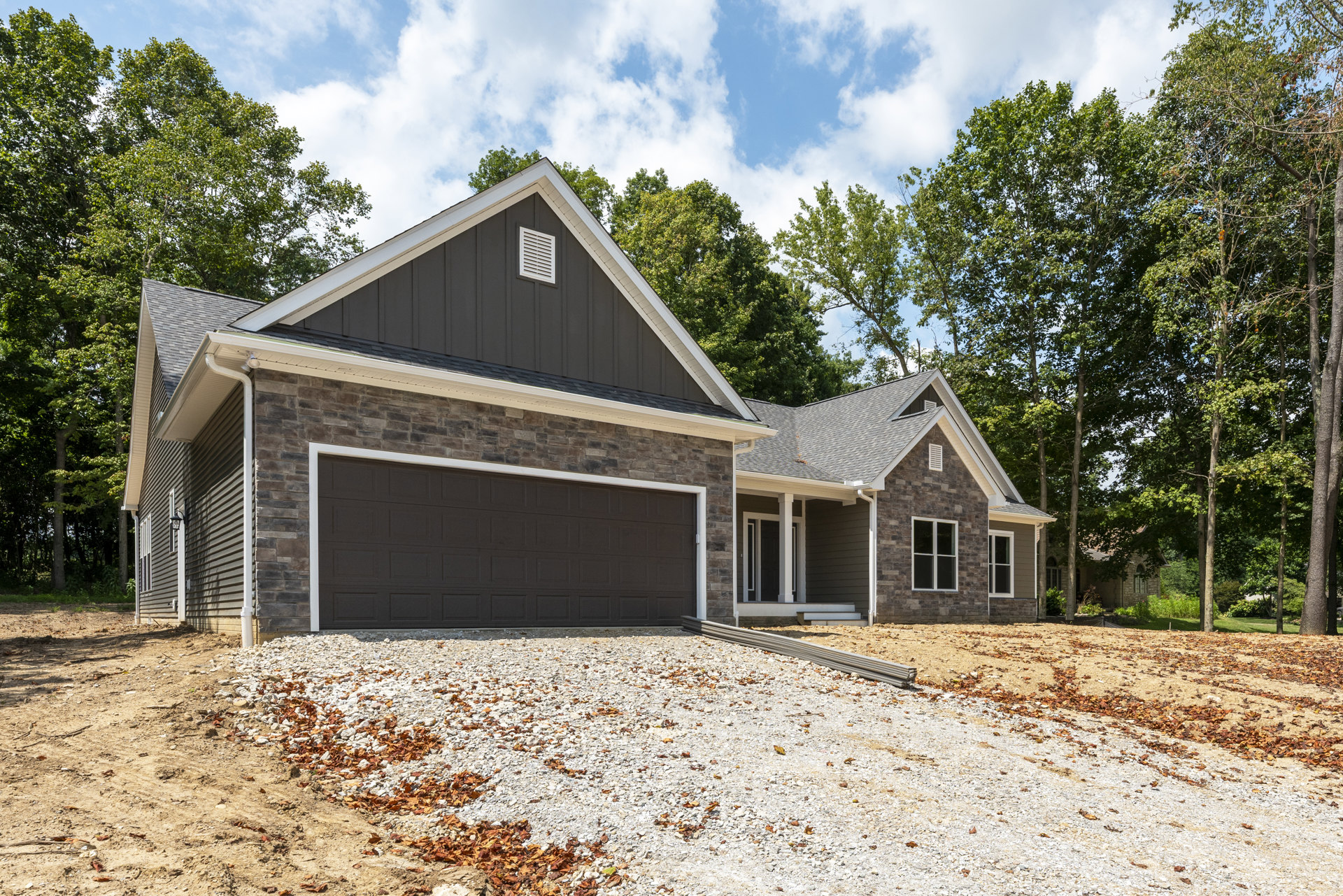 Modern house with black siding, white-framed windows, brick garage wall, gravel driveway scattered with leaves, surrounded by trees under a cloudy sky