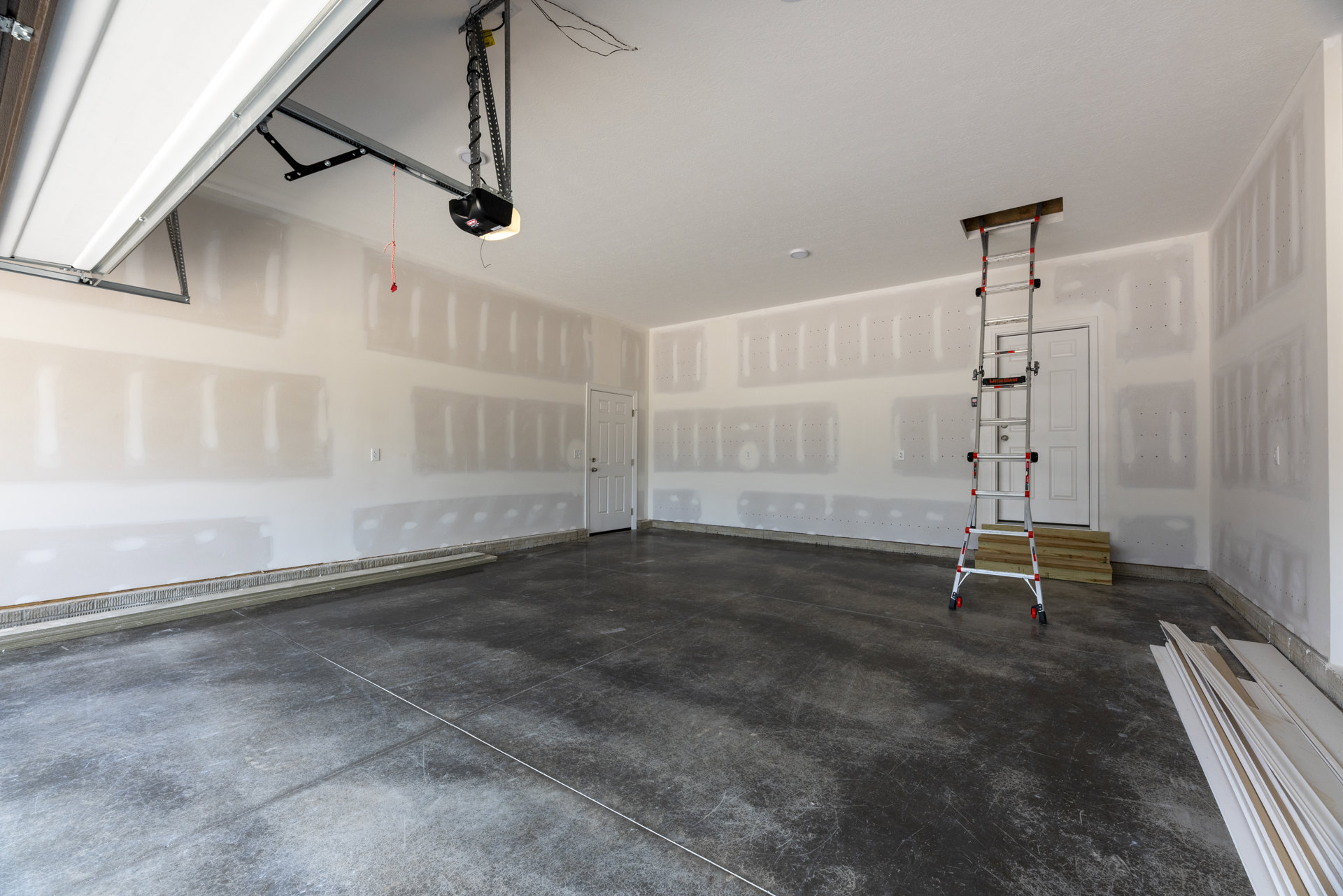 Garage interior with white plaster walls, a white door with silver knob, wood boards and a whiteboard on the floor, and a metal ladder leaning against the wall