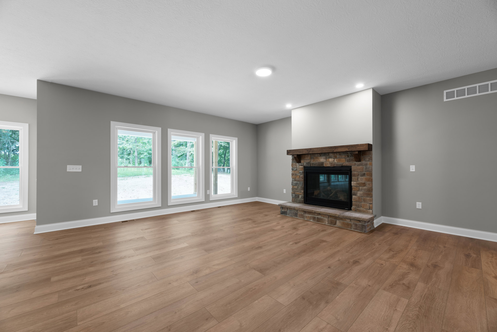 Hardwood floor living room featuring a central fireplace with glass doors and a wood mantel, large windows framing views of leafy trees outside