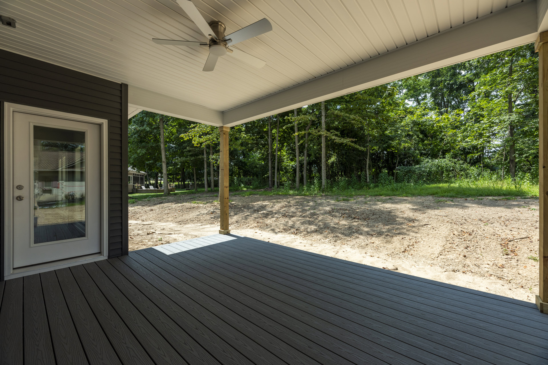 Covered deck with four-blade ceiling fan, wooden post featuring a hole, glass door reflecting nearby house, surrounded by leafy trees in the background