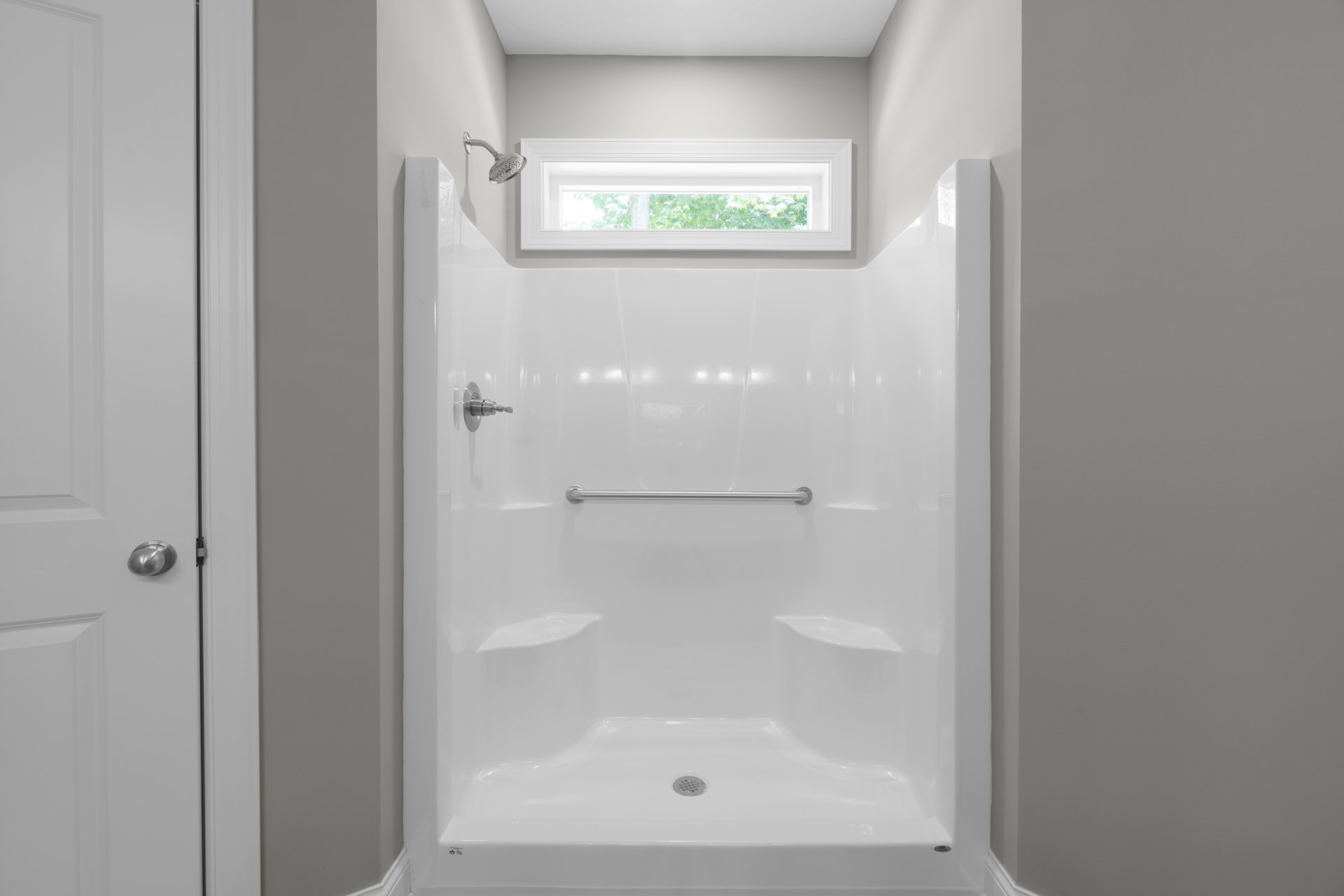 White-tiled shower with glass door, chrome shower head, window with white frame, and nearby door handle and light switch on wall