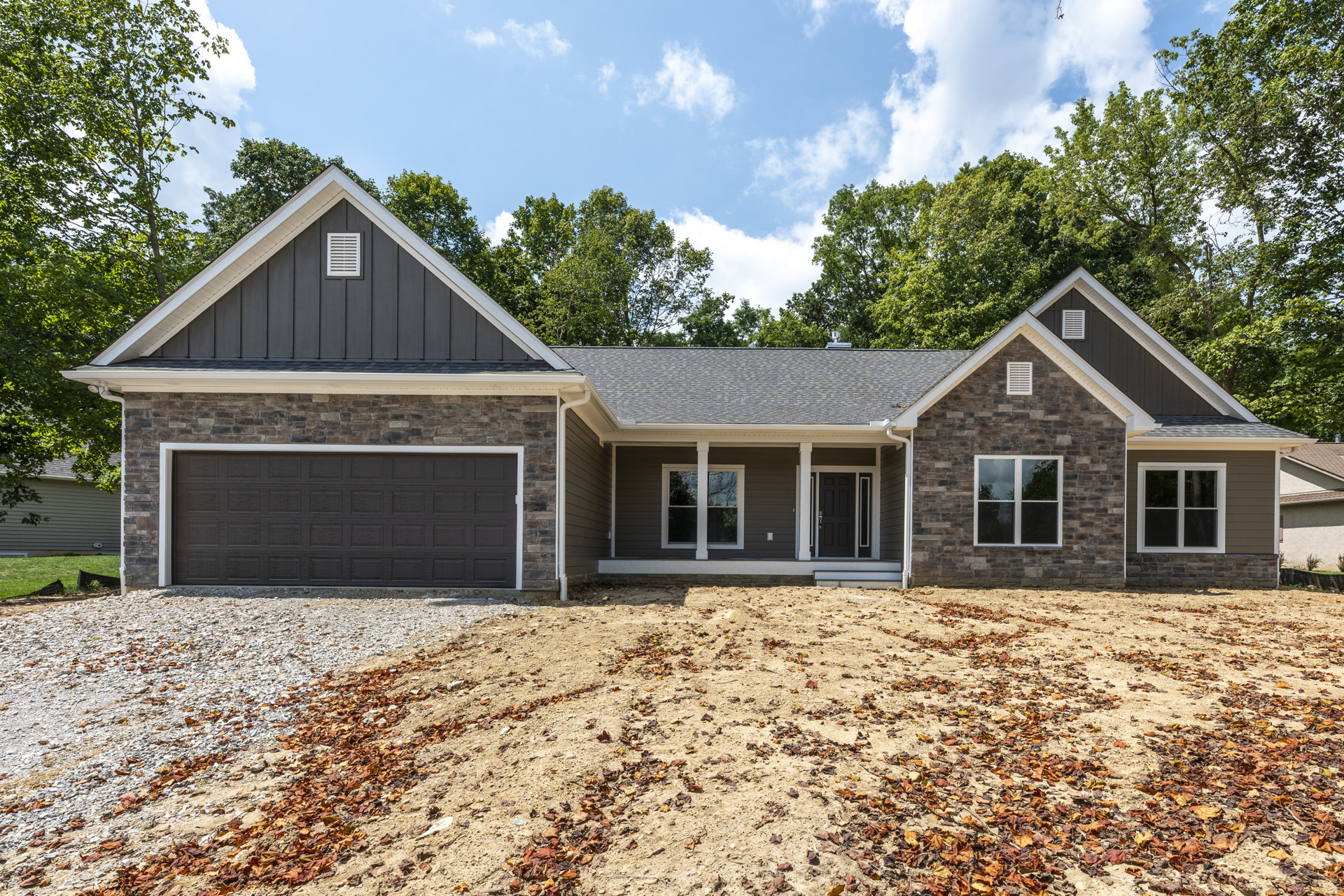 Brick and stone exterior home with white-framed windows, attached garage door, paved driveway bordered by dirt and scattered leaves, mature trees in the background.