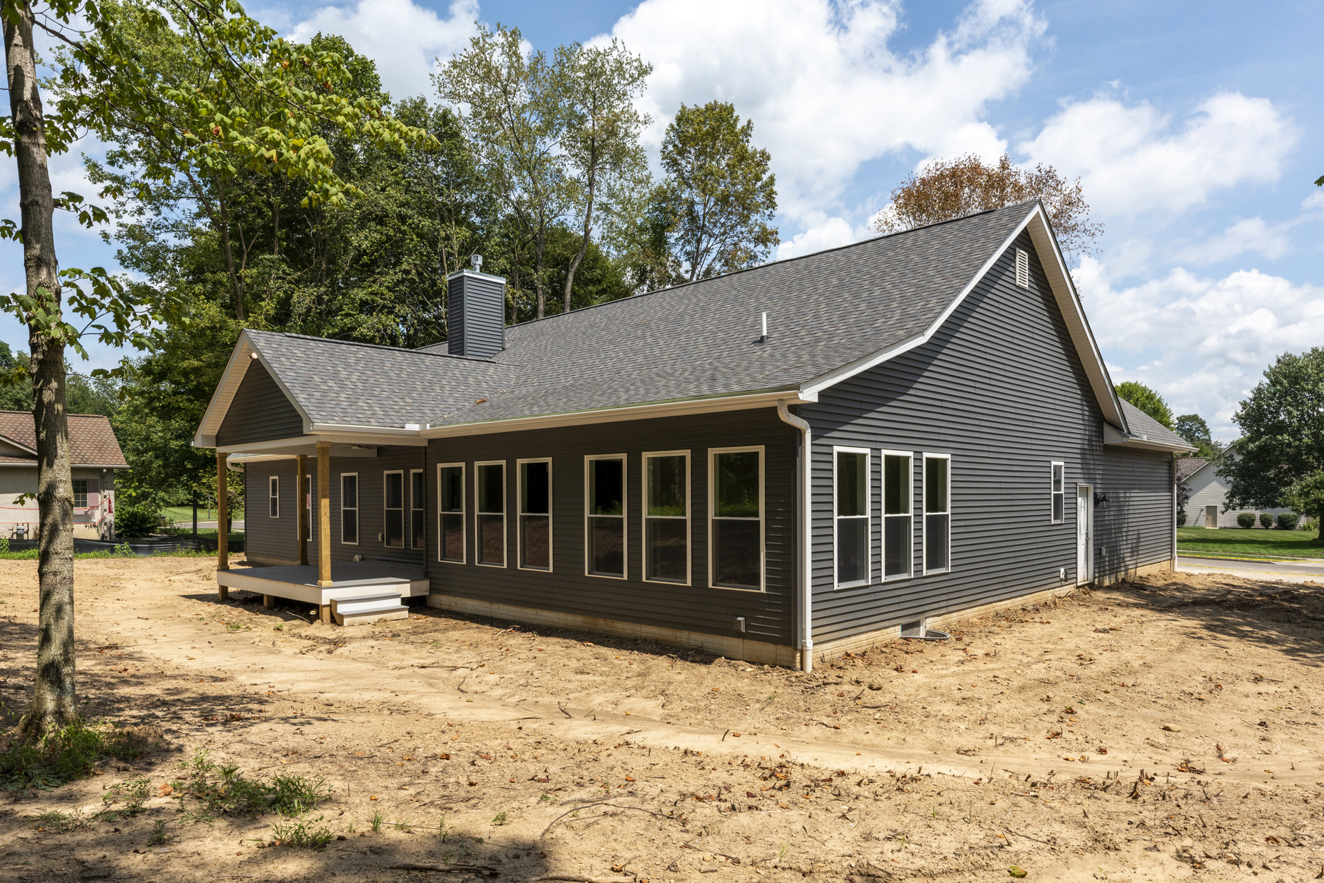 Partially built house with exposed wooden framing, multiple white-trimmed windows, unfinished porch supported by wooden posts, dirt path alongside, surrounded by tall trees under