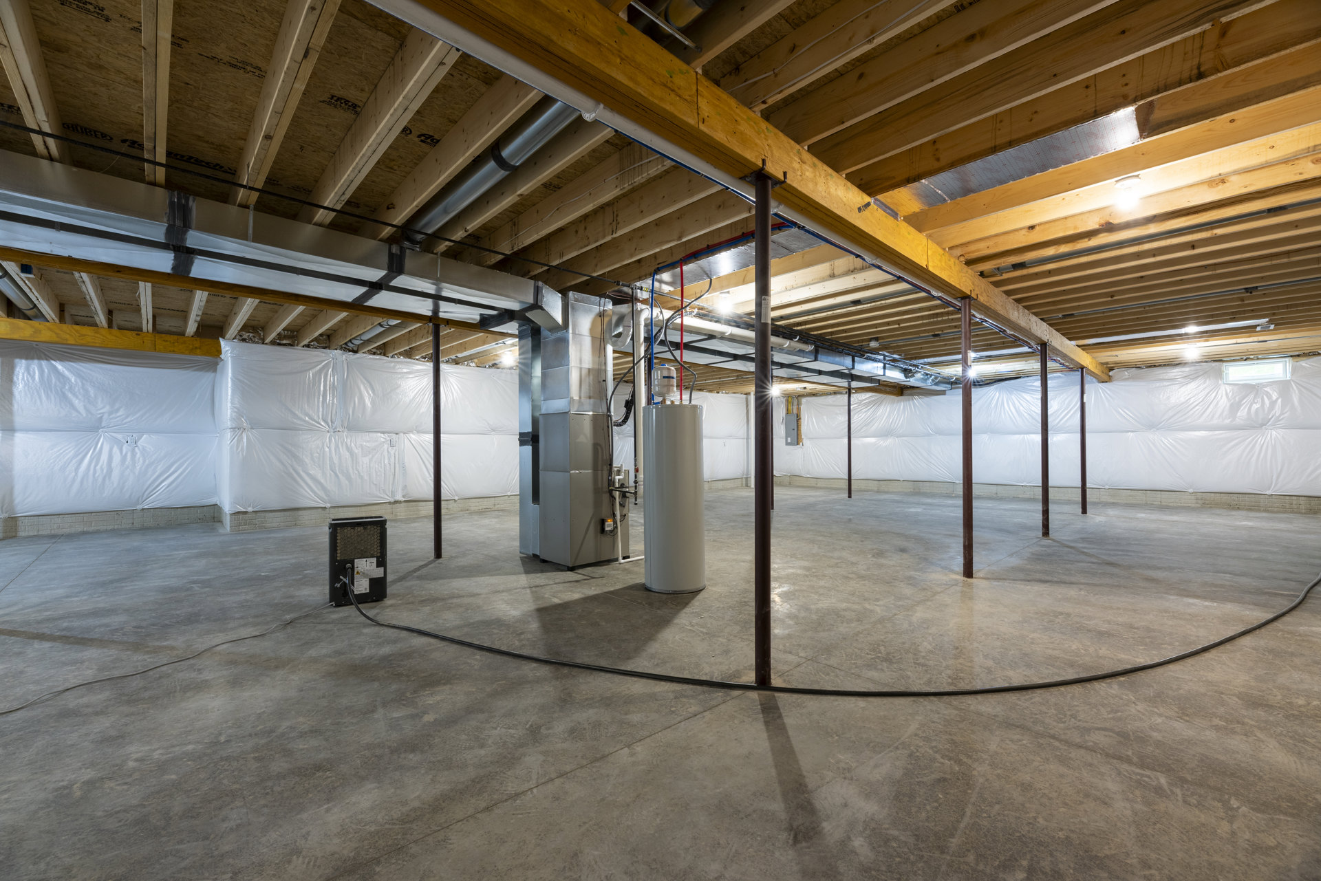 Utility room with exposed steel beams, composite flooring, white cylindrical water heater featuring a red button, and visible plumbing pipes along the ceiling and walls