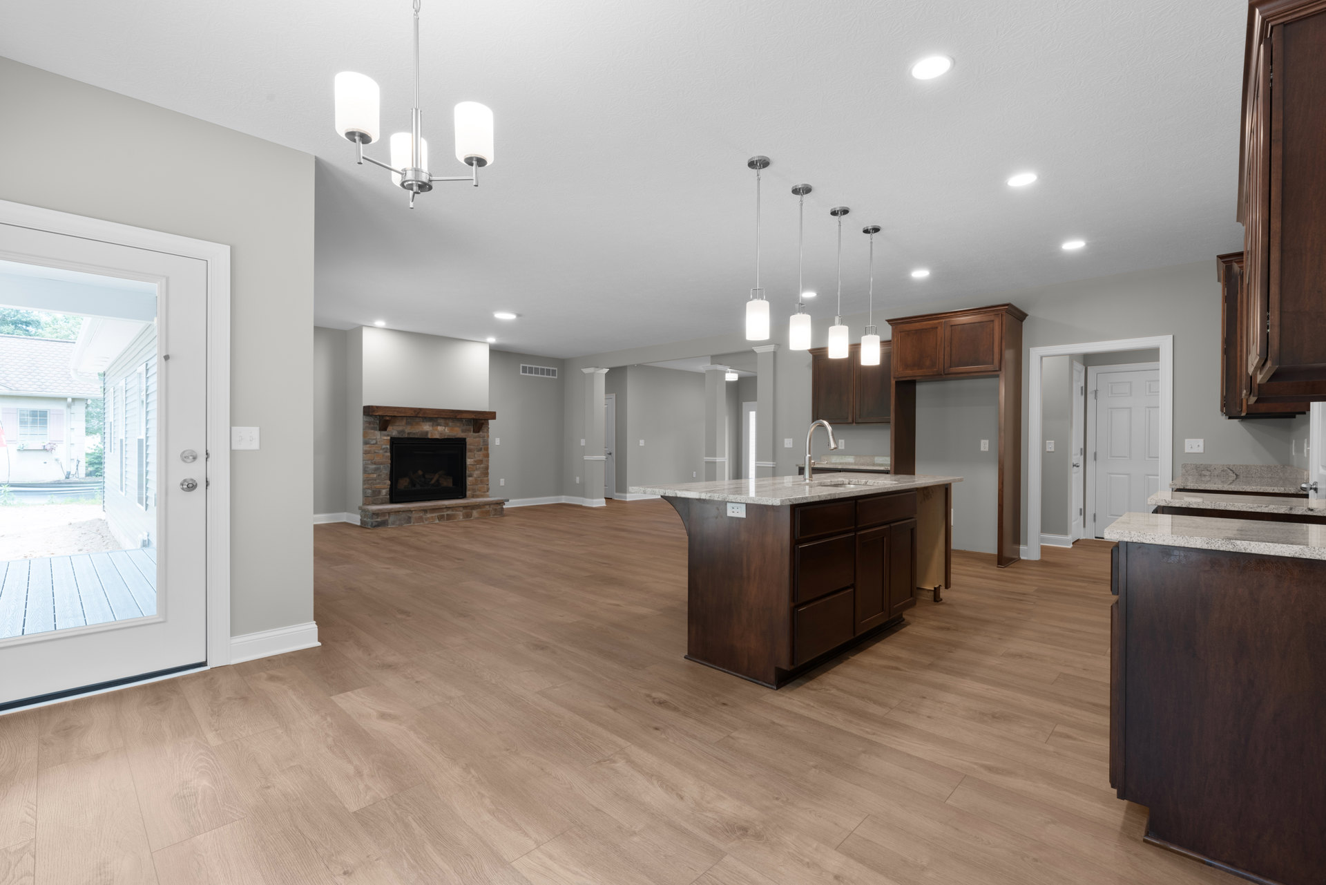 Spacious open floor plan featuring a central kitchen island with sink, modern cabinetry, wood flooring, built-in fireplace with stacked logs, ceiling light fixture, and adjacent