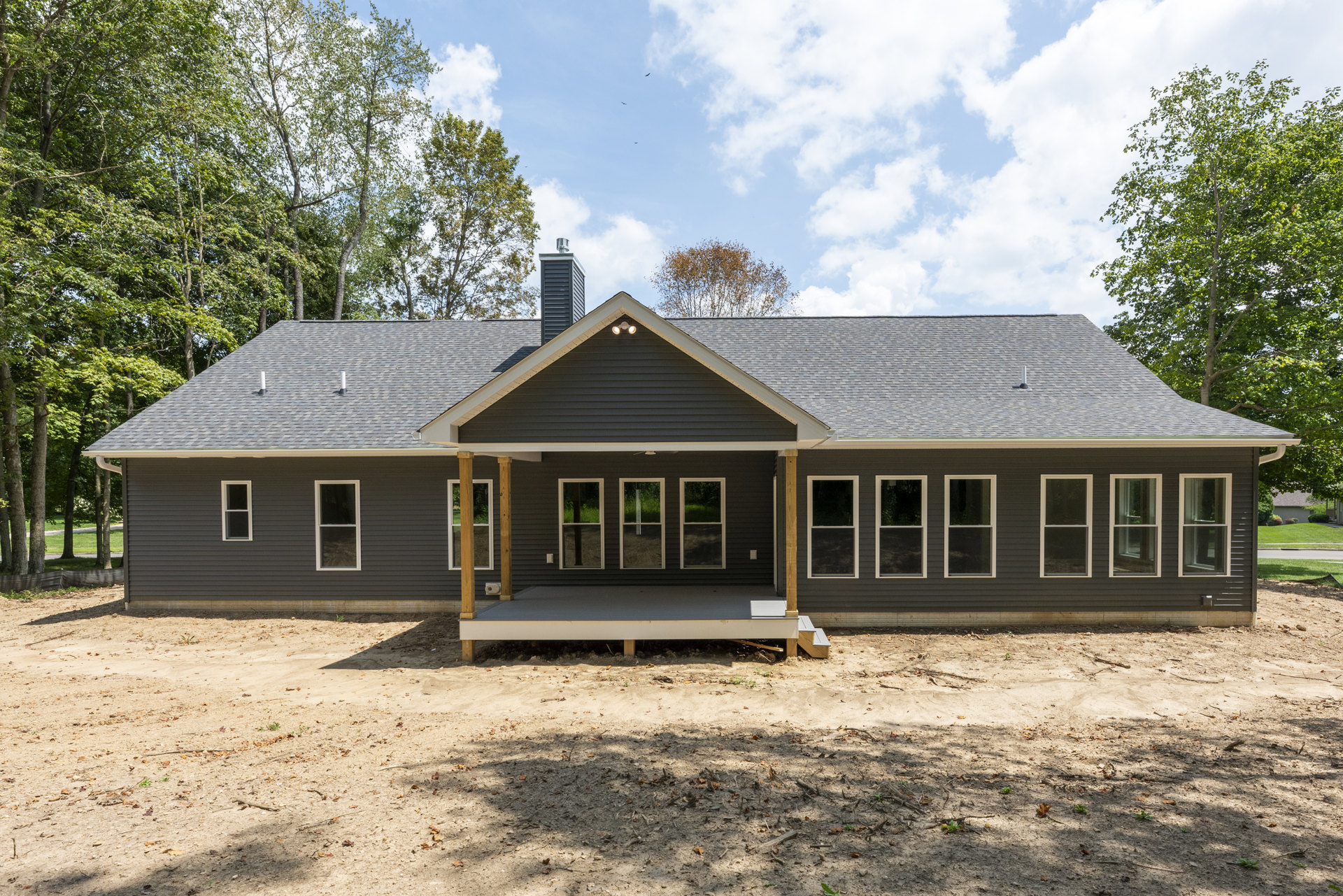 Front porch with white railing, dirt yard, stairs leading to entry, white-framed windows, gabled roof, small white cottage visible in background, scattered trees under partly