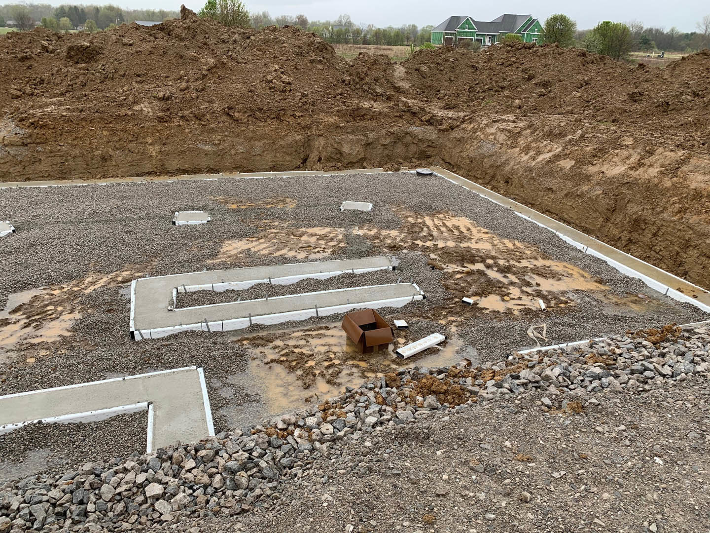 Construction site with exposed soil and rocks, green house with white door in background, brown utility box with opening, white rectangular object on ground, concrete drainage