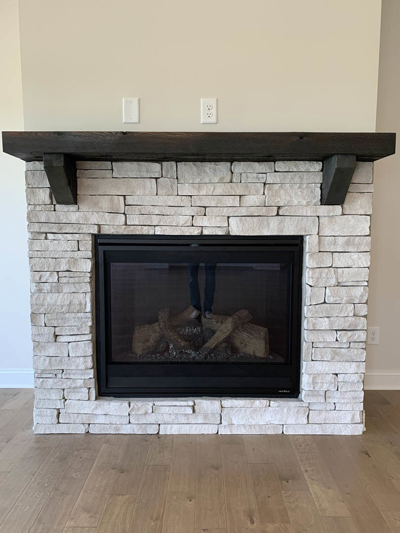 Stone fireplace with wood mantle, fire screen, and wood floor; person wearing blue jeans rests feet on tree stump in front of hearth; stone wall and indoor room details visible.