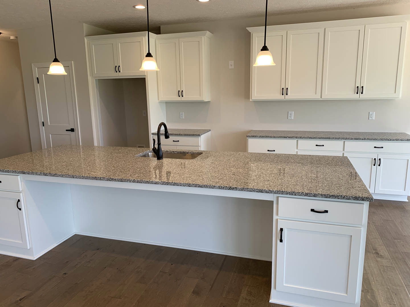 White kitchen with granite countertops, central island featuring a sink and black faucet, white cabinets with black handles, tile backsplash, and hardwood flooring.