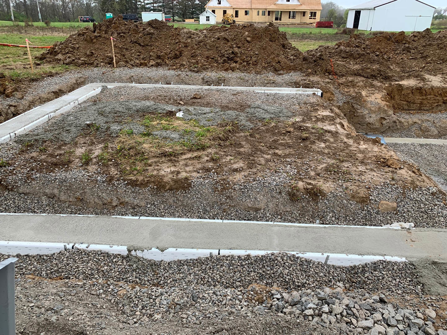 Dirt and gravel foreground leading to a modern house with multiple windows, surrounded by sparse grass and trees under a clear sky