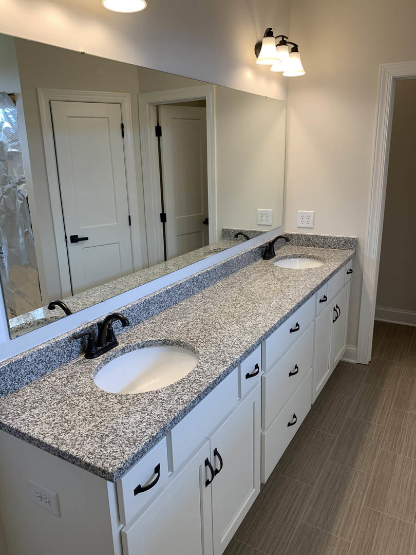 Bathroom with double white sinks, wide mirror above, light fixture with three bulbs, white cabinetry, tile backsplash, and wood flooring.
