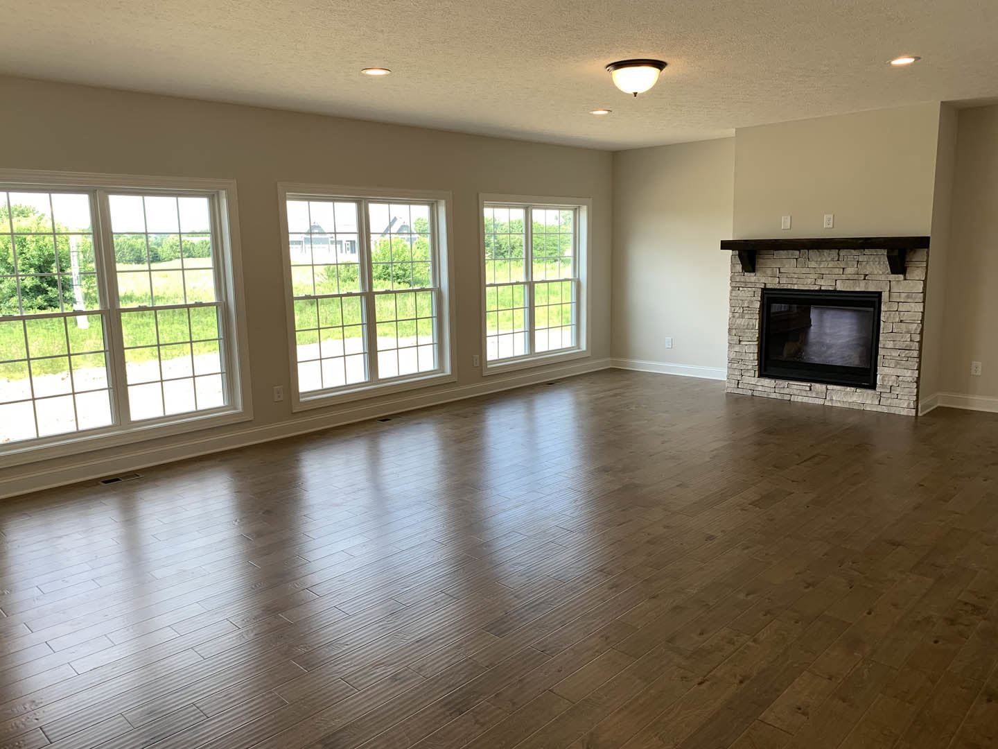 Living room with wood flooring, large multi-pane windows, and a fireplace featuring a black screen; windows offer a view of a neighboring house.