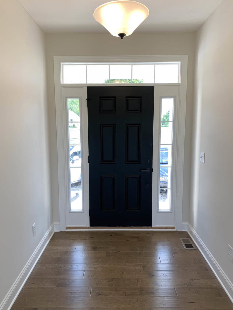 Black door with white trim in a hallway featuring hardwood floors with white baseboards, ceiling light fixture, and window with white shade