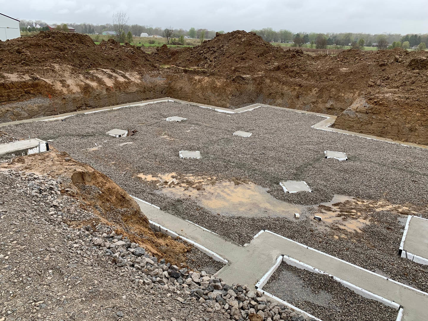 White house with black roof surrounded by dirt and gravel piles at construction site, leafless tree nearby, overcast sky above
