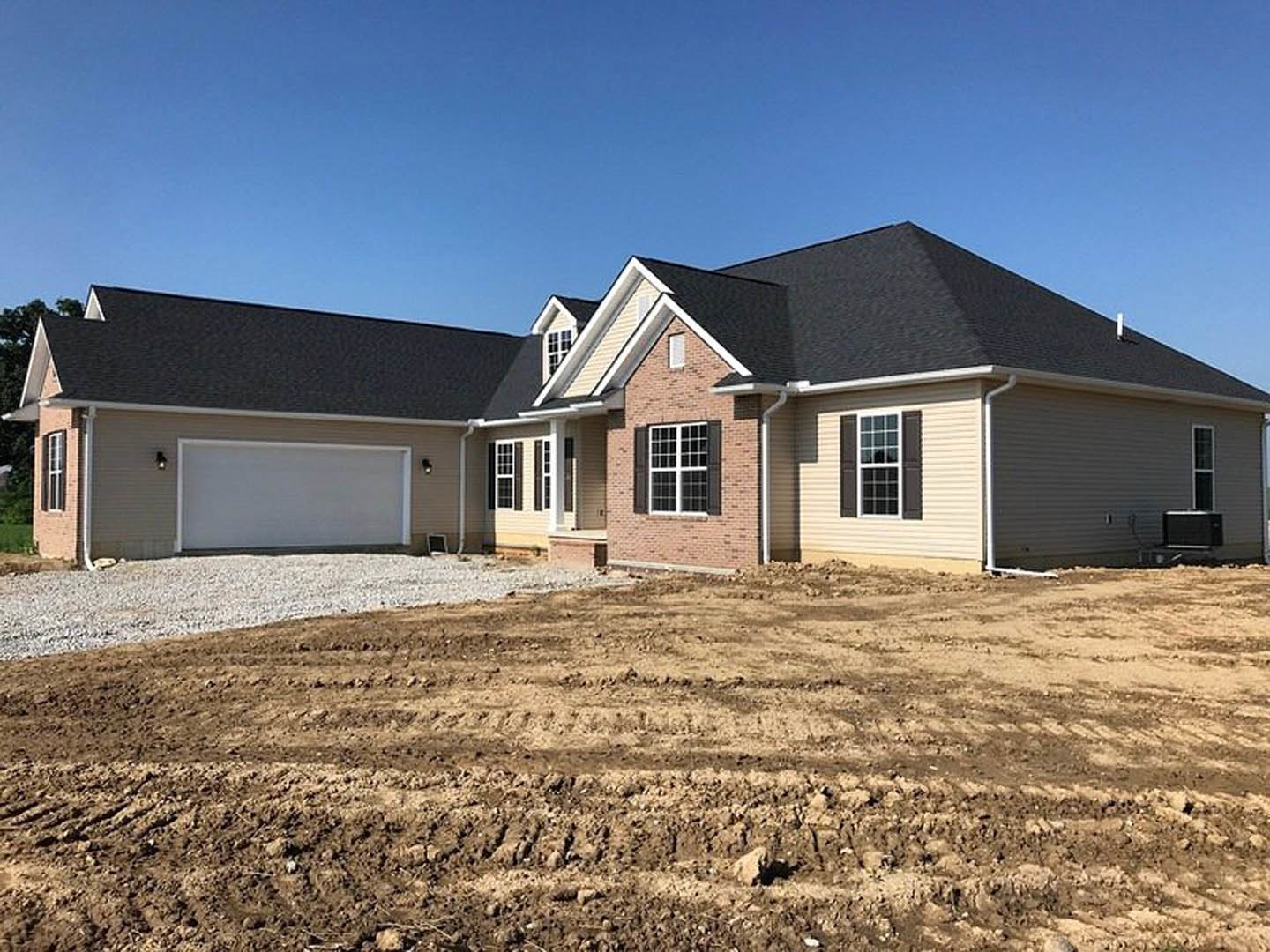 White siding house with black roof under construction, dirt patch and tire tracks in foreground, white-framed window, gravel and grass along foundation.