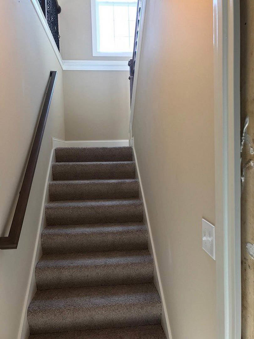 Carpeted staircase with white walls, wooden handrail, and window allowing natural light into the interior hallway
