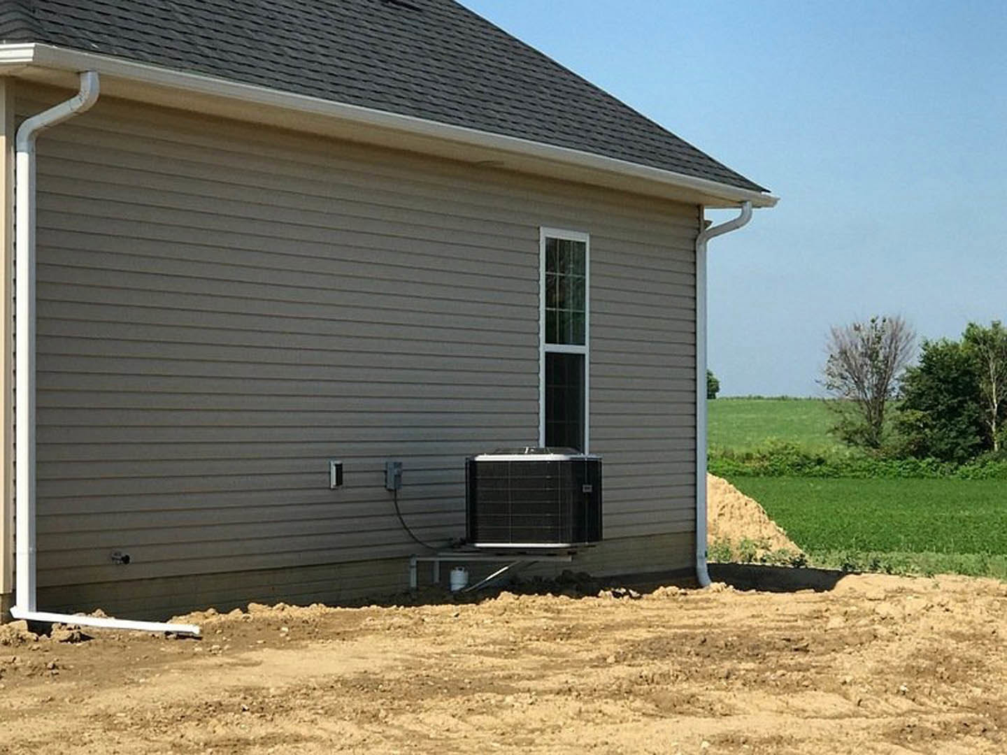 Two-story home with light-colored siding, white-framed windows, large black and white heat pump unit beside the house, leafless tree, dirt yard, and white barrel in foreground.